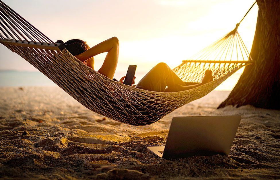 Persona desconectando en vacaciones frente al mar, aunque revisa el móvil desde la hamaca